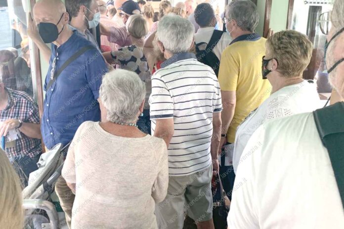 Vaporetti di Venezia oggi. Due anziani residenti si tengono per mano accingendosi ad attraversare la marea umana di turisti per scendere